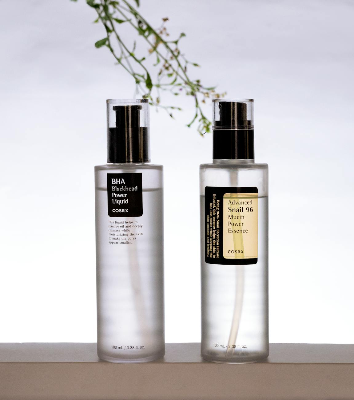 Two COSRX skincare bottles. Left: BHA Blackhead Power Liquid. Text: This liquid helps to remove oil and deeply cleanses while moisturizing the skin to make the pores appear smaller. 100 mL / 3.38 fl. oz. Right: Advanced Snail 96 Mucin Power Essence. Text: Being 96% Snail Secretion Filtrate (Mucin), this essence helps the skin to lose less moist while keeping the skin smooth and healthy. 100 mL / 3.38 fl. oz.